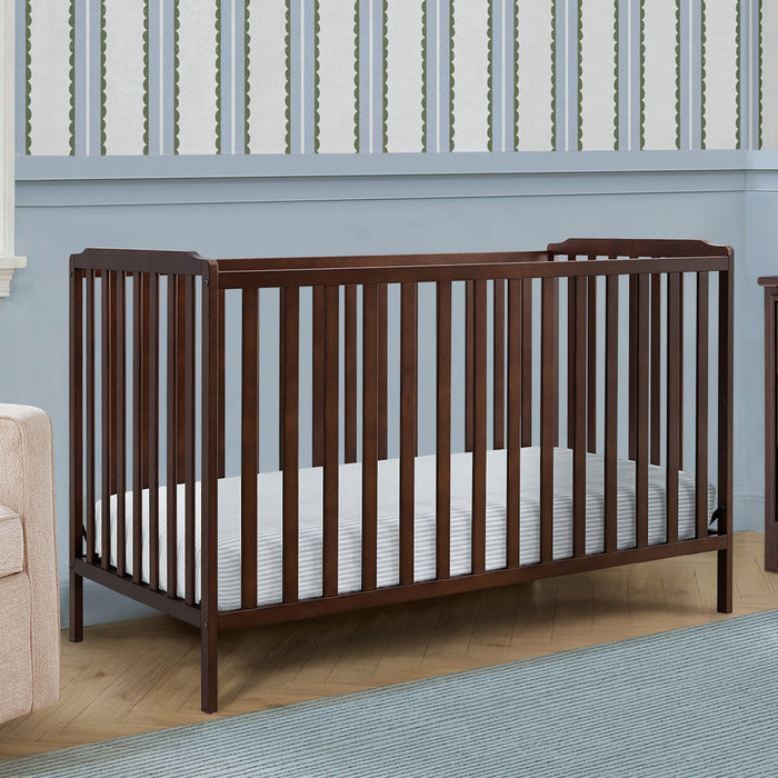 Wooden crib with white mattress in a room with light blue walls and wooden flooring. 1-Emmie 4-in-1 Convertible Crib