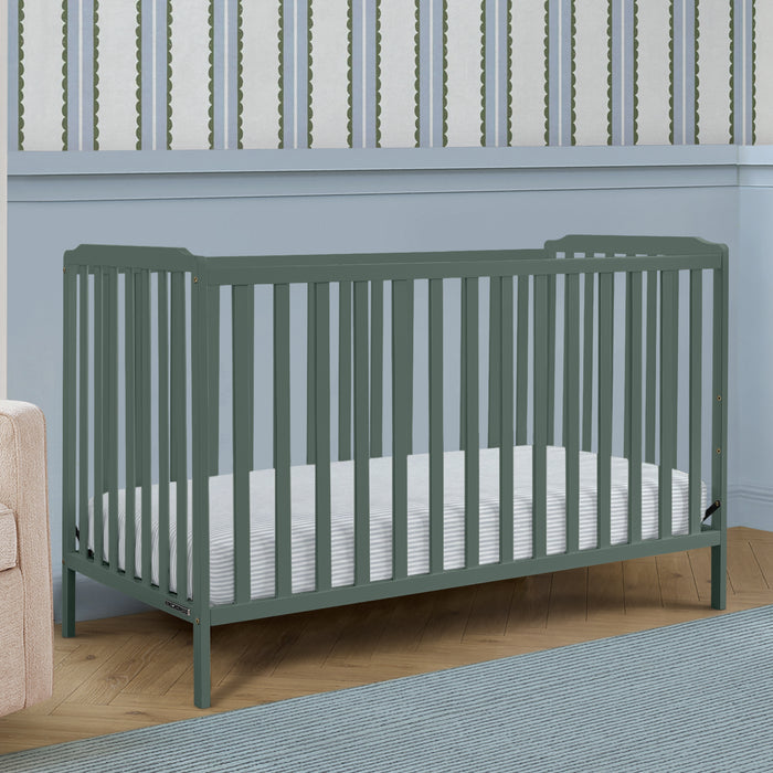 A sage green convertible crib with vertical slats, shown with a white mattress in a styled nursery setting with a light blue wall, wood floor, and rug. 1-Emmie 4-in-1 Convertible Crib