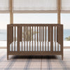 Wooden crib with white mattress in a room with large windows and plaid rug 24-Jalen Grow With Me 9-in-1 Convertible Bassinet to Crib