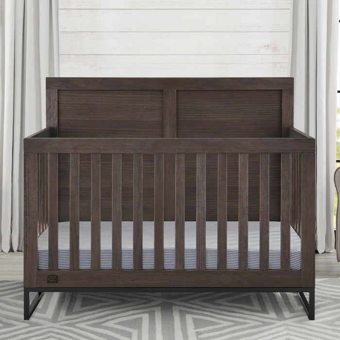 Wooden crib in a room with light-colored walls and a geometric-patterned rug. 3-Foundry 6-in-1 Convertible Crib