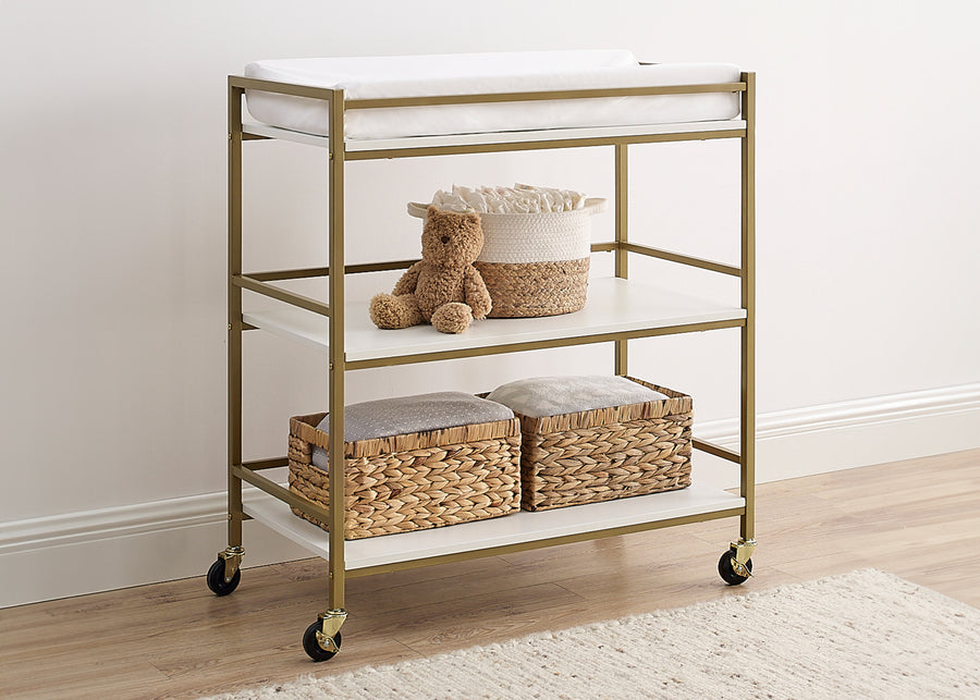 Gold-framed rolling cart with white top and shelves, containing a teddy bear and woven baskets on a wooden floor. 4-Jade Changing Table