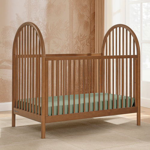 Wooden crib with a green mattress in a room with patterned wallpaper and a window.