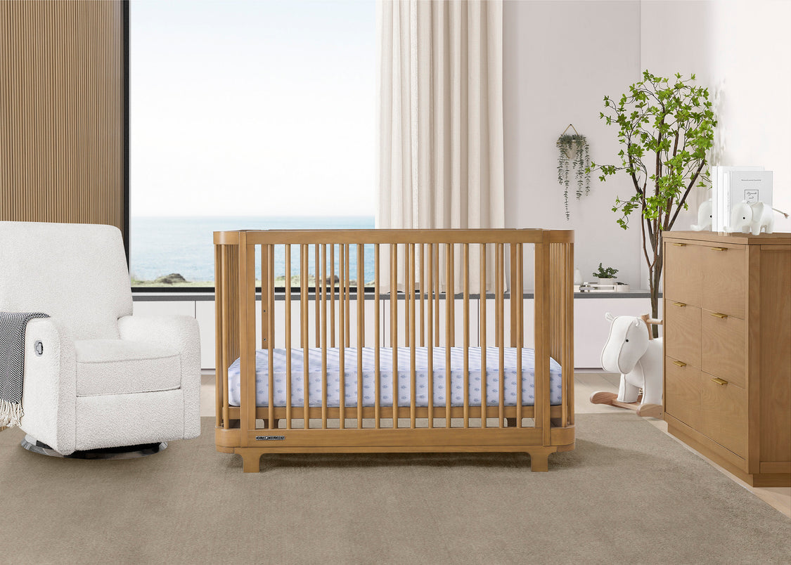 A brown Nest 4-in-1 Convertible Crib by Delta Children in a nursery room, with a white chair and a plant in the background.
