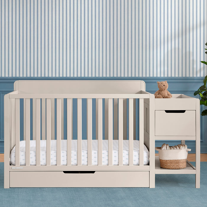 Beige crib with white bedding in a room with striped wallpaper 2-Maya 5-in-1 Convertible Crib and Changer with Storage