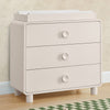 Beige dresser with three drawers on a wooden floor. 27-Ari 3 Drawer Dresser with Changing Top