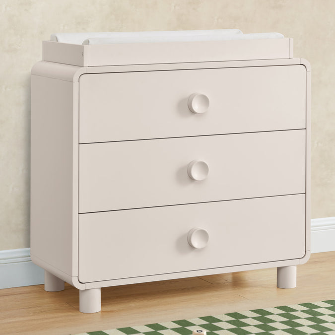 Beige dresser with three drawers on a wooden floor. 27-Ari 3 Drawer Dresser with Changing Top