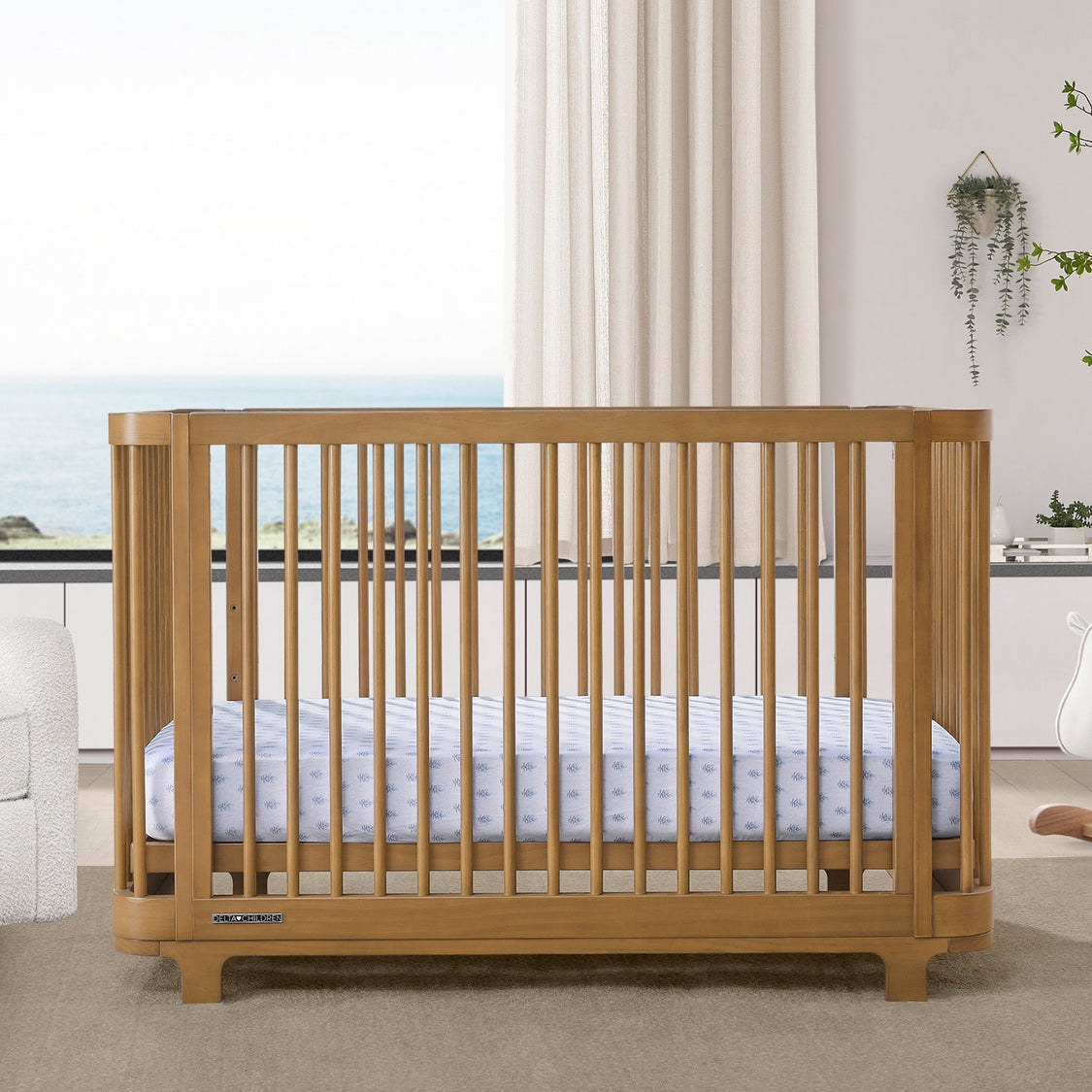 A brown Nest 4-in-1 Convertible Crib by Delta Children in a modern nursery room, with a white mattress on a white floor, alongside a white chair and a wooden dresser.
