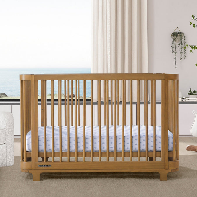A brown Nest 4-in-1 Convertible Crib by Delta Children in a modern nursery room, with a white mattress on a white floor, alongside a white chair and a wooden dresser. 30-Nest 4-in-1 Convertible Crib