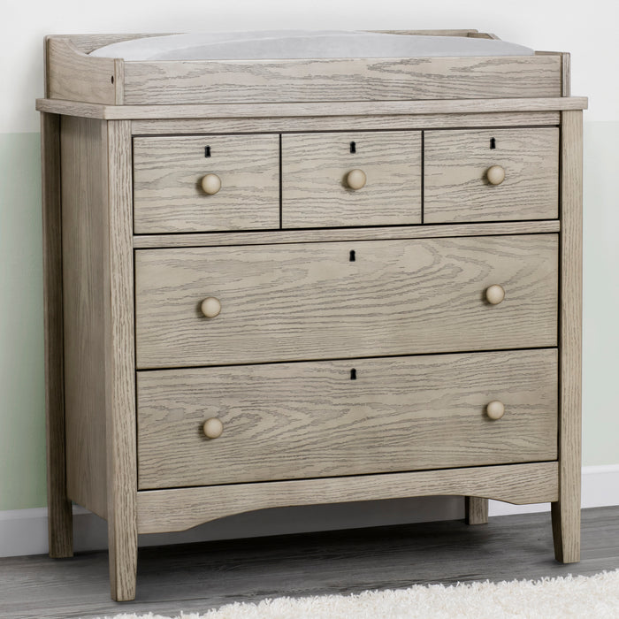 A black 3 drawer dresser with a changing top, set on a room with a striped curtain, a teddy bear, and a plush toy. The dresser has a rustic design with wood drawer pulls and a lightly textured finish. 3-Farmhouse 3 Drawer Dresser with Changing Top and Interlocking Drawers