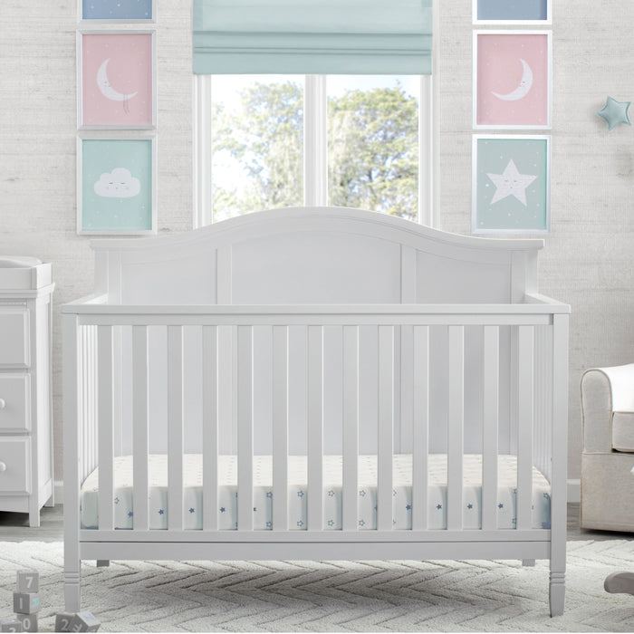 A white 5-in-1 convertible crib with a headboard, situated in a nursery room with decorative wall art, curtains, and furniture. 3-Madrid 5-in-1 Convertible Crib